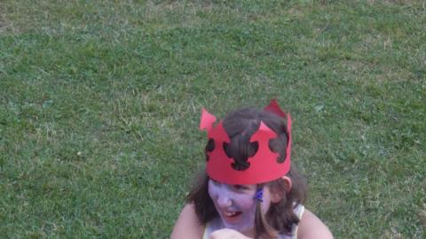 Camper with painted face and paper crown braiding the very short hair of another camper and laughing.