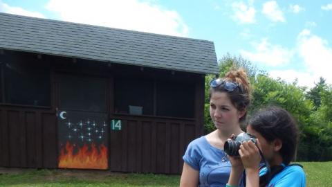 Camper looks through camera at something out of frame with counselor looking at target also near camper.