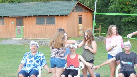 Staff seated in lawn chairs as cabin group covers their head in shower cap and shaving cream.