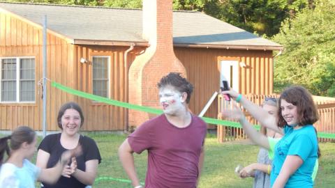 Smiling cabin group presents counselor who has been made over with thick facial make-up posing for the rest of camp. 