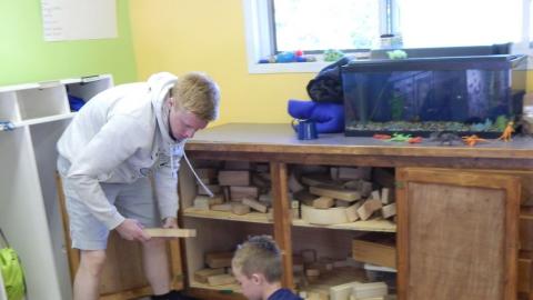 Youth playing blocks on the floor with counselor standing at block cupboard handing him a block.