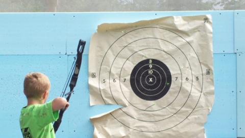 youth draws his bow to shoot at a paper target in archery class.