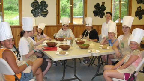 Chef's Club participants smiling at the camera wearing paper chef hats as they enjoy a salad they've made.