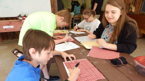 Youth sit with counselor at a table drawing 9 man morris boards on cloth with sharpie markers.
