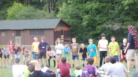 Two cabin groups collaborating on a sit. Everyone in a line looks to their left at a counselor wearing a towel on their head.
