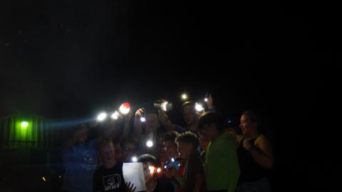 cabin group gathered together in the dark with flashlights to help them sing lyrics written on papers they are holding.