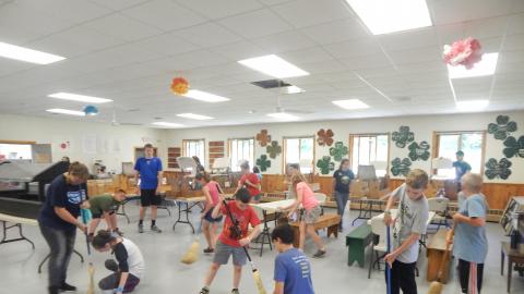 Campers working together to sweep the dining hall floor. Some youth are kneeling with dustpans as the others sweep.