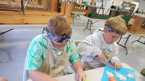 Two young men sit at a table cutting carrots and onions.
