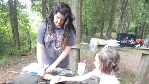 Two youth add bread to pie iron cookers outdoors. One is wearing a princess crown.