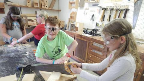 One youth holds a board while another youth cuts it with a hand saw.