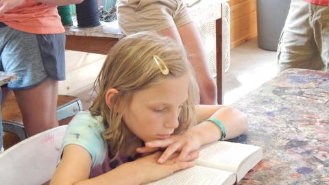 Youth sits at table with head resting in hands as they read a book.