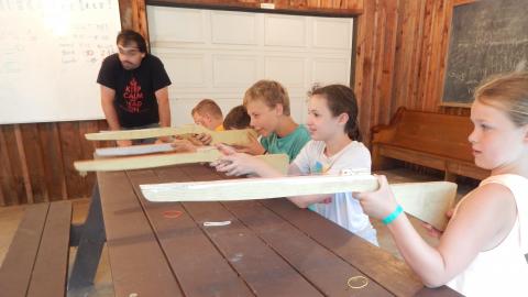 Youth seated at a picnic table aim wooden rubber band rifles as a counselor looks downrange.