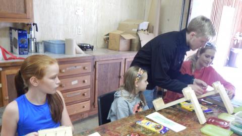 Counselor leans over to help camper working on wooden loom. Neighboring campers watch.