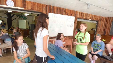 Two youth on opposite sides of a table smiling at one another while others look on.