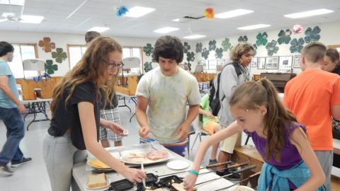 Campers gathered around a table add bread, cheese, and meat t a pie iron cooker.