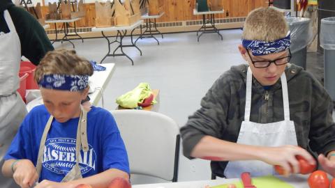 Two youth seated at a table cutting up tomatoes and peaches.