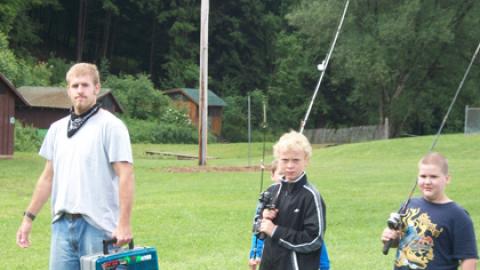 counselor with tacklebox leads group of youth with fishing poles across camp toward the pond to fish.
