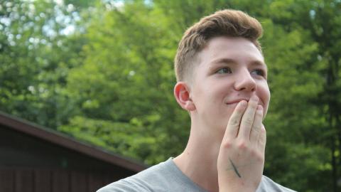 Youth with hand to face smiling and looking away from the camera with cabin and tree in background.