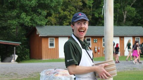 Kitchen assistant looking into camera and smiling as they carry a stack of plates outside. 