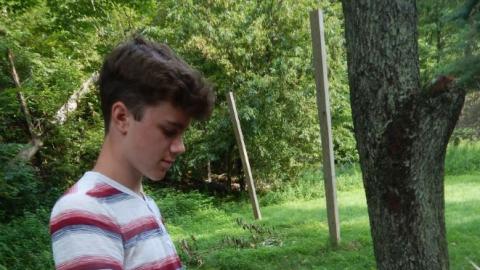 Youth standing in wooded area whittling a stick with a knife.