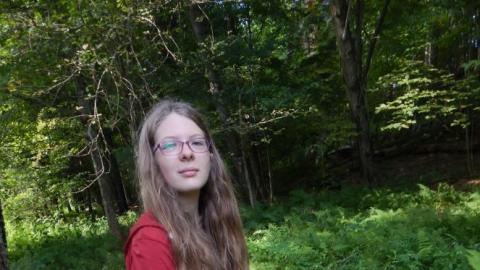 Youth standing in wooded area whittling stick with a knife.