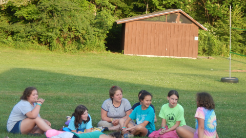 Cabin group seated together on the ground talking and laughing while the counselor "sleeps" facedown on the ground in the center of their circle.