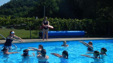 Lifeguard looks over pool as Aquatics Director demonstrates a swim stroke where arms are extended in front. Campers copy from within the pool.