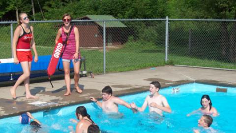 Campers in the swimming pool looking at two lifeguards standing over them on the pool deck laughing.