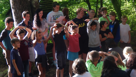 Platoon group performing a skit for the rest of camp. Youth in front row have hands raised over their heads with hands together as a counselor reads from a paper.