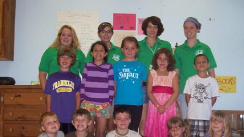 Group of day campers with staff in back holding wooden sign made of sticks that reads "Day Camp"