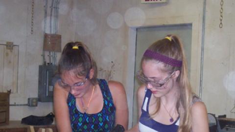 two teen girls working together to hold pieces of a chair in place while the other holds a drill.