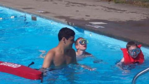 Lifeguard with two campers near pool stairs. One is wearing a life jacket. Both are wearing goggles.