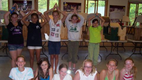Two rows of youth. One row standing in back with hands raised above head. One group in front on floor in upward dog pose.