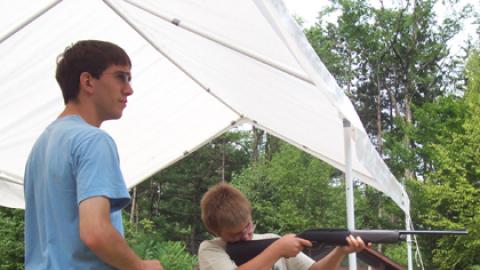 Counselor looks on as youth takes aim with air rifle outdoors.