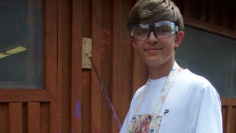 Camper wearing paint covered apron and goggles behind wood shop dipping brush into can of paint.