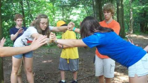 Two youth on opposite cables balancing against each other a few inches from the ground while other campers spot from behind. 