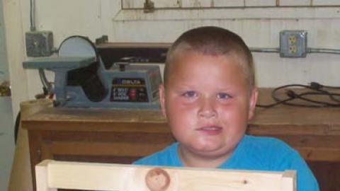 Youth stands behind a wood project with a smirk on face. 