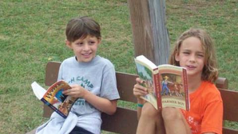 two youth sitting together on a bench reading books.