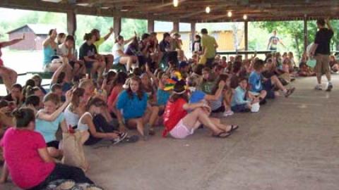 Campers seated in rec hall with hands raised.