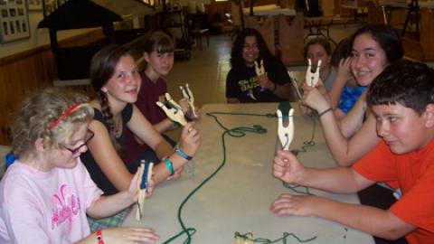 youth sitting around a table, looking at the camera and showing the process of lucetting with wooden tool and yarn.