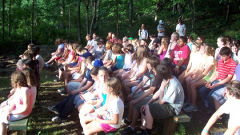 campers seated on benches in a wooded area watching a performance by a platoon group in front.