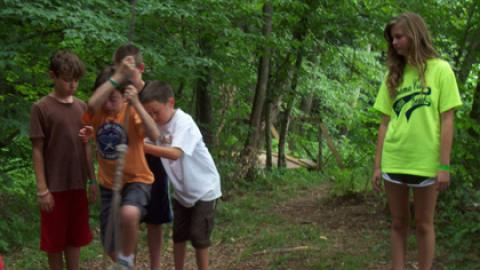 Youth puts foot up into rope from wooden platform. Three campers on platform assist.