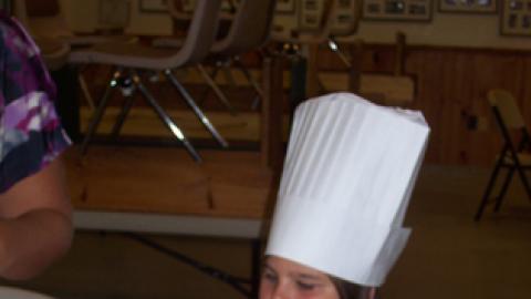 Youth wearing paper chef hat spooning batter into muffin liners.