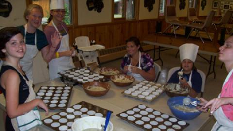 Youth around a table spooning batter into muffin tins while wearing aprons, chef hats, or hair nets.