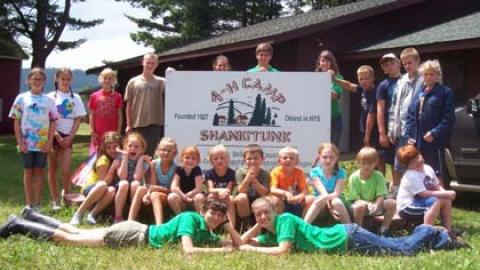 group of day campers standing around and sitting in front of 4-H Camp sign with counselors on the ground in front of them.
