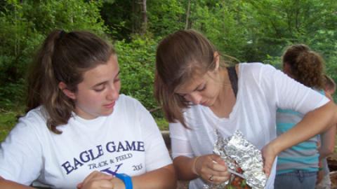 Cookery staff dump contents of hot foil packet onto paper plate for camper.
