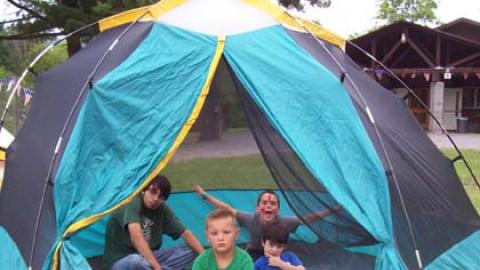 Youth in a large tent with counselor. One has arms raised and face painted yelling at camera. One sits cross legged. One sides to the side stroking chin. One lays on back.