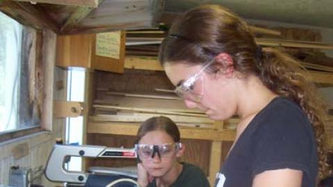Staff using jig saw to assist a camper with a wood project as the camper looks on.