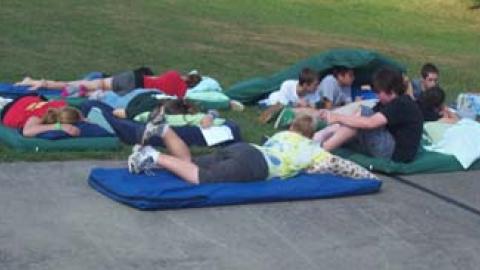 youth and counselors rest outside their cabin on mattresses on a beautiful day.