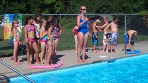 Youth stand at poolside with pool noodles getting ready for a relay race under the supervision of a lifeguard.